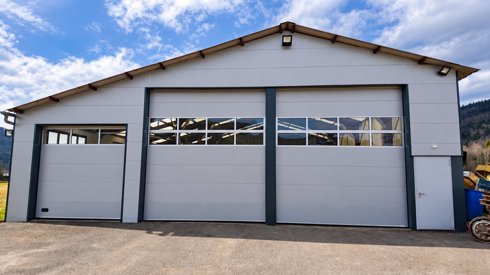 Façade d'un hangar gris à bardage métallique équipé de trois grandes portes sectionnelles dont deux avec bandeaux vitrés, et d'une porte piétonne, sous un ciel nuageux.