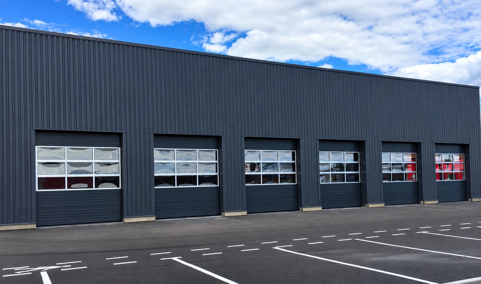 Façade d'un bâtiment industriel en bardage gris foncé équipé de six portes sectionnelles vitrées, devant un parking aux marquages blancs.