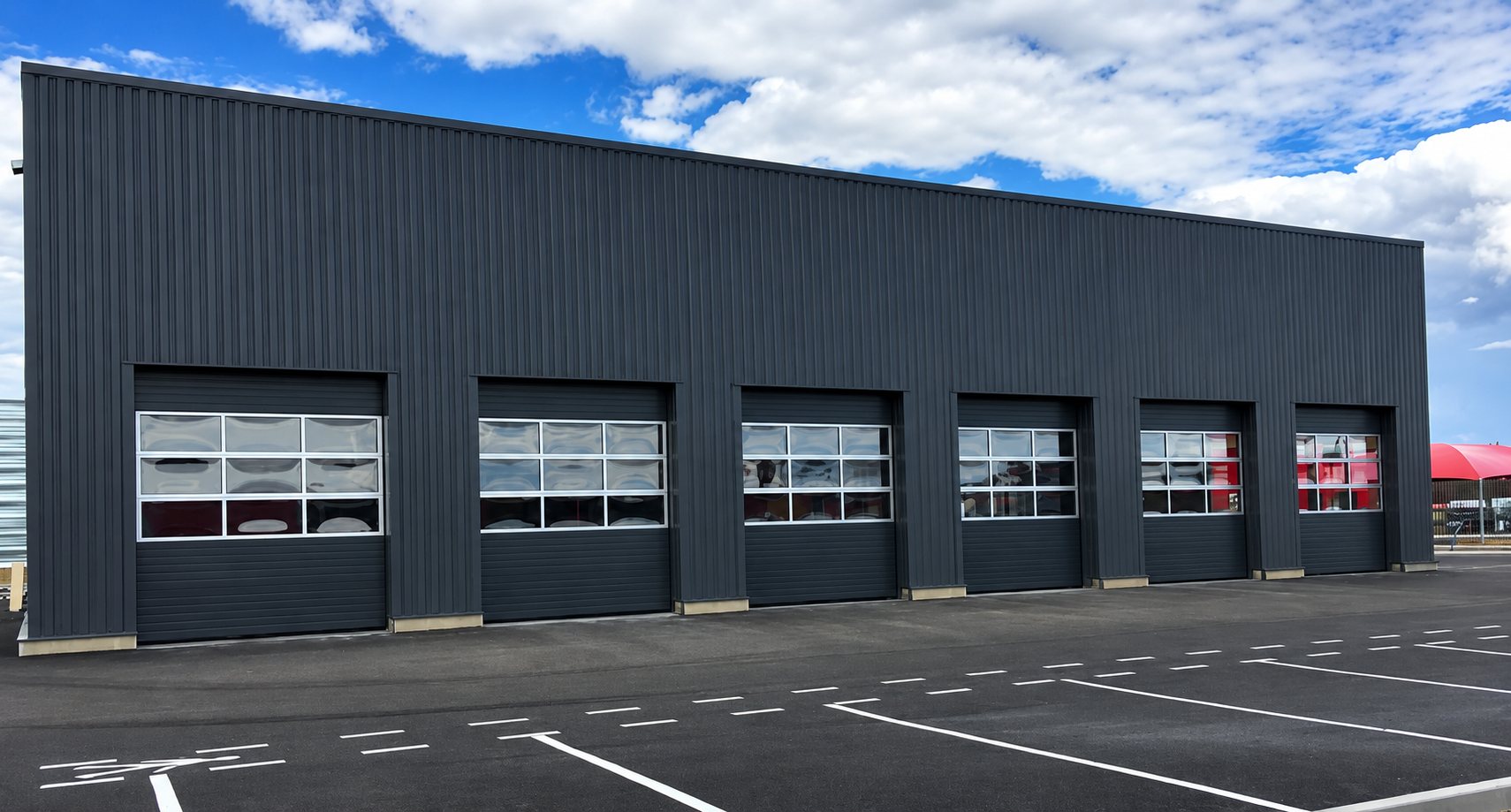 Façade d'un grand bâtiment métallique gris foncé équipé de six portes sectionnelles à hublots vitrés, devant un parking aux places tracées en blanc.