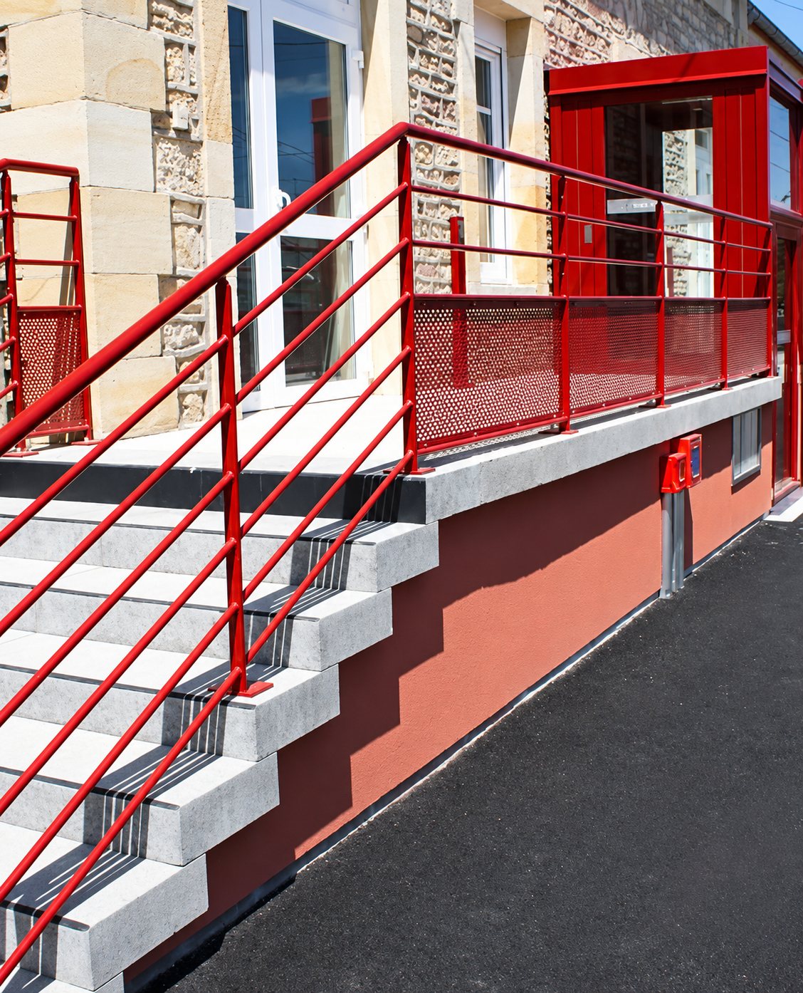 Un escalier en béton et une rampe d'accès bordés d'un garde-corps métallique rouge, devant une façade en pierre et un mur rouge.