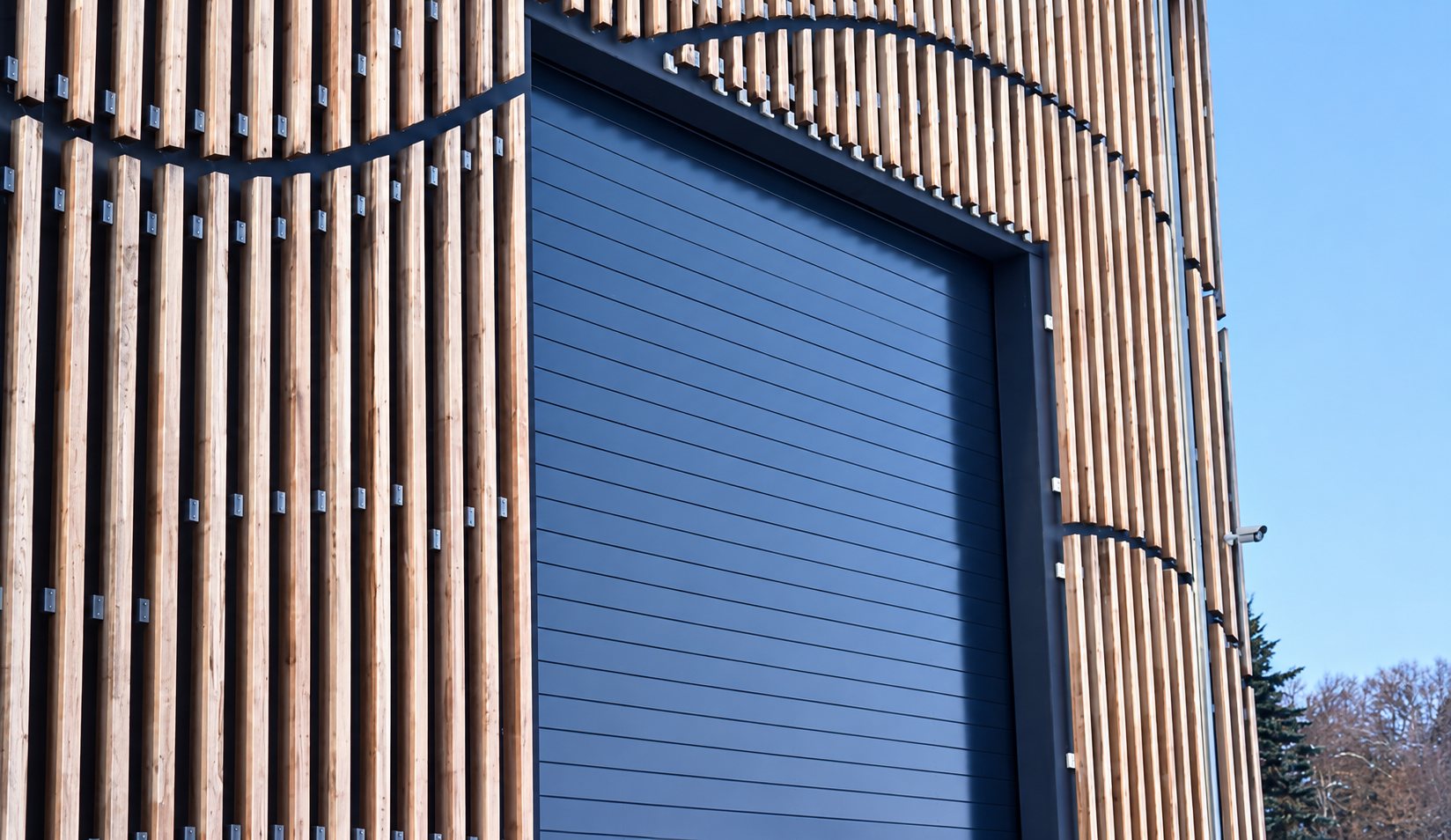 Façade d'un bâtiment habillée de lames de bois verticales courbes autour d'une grande porte sectionnelle gris foncé, avec des arbres en arrière-plan.
