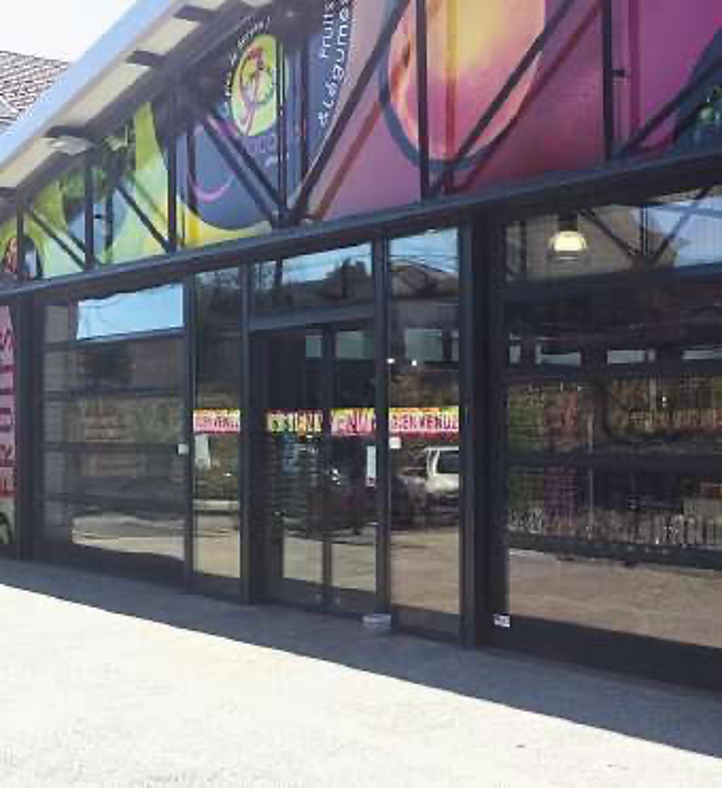Devant d'un magasin aux grandes vitrines noires, surmonté de grandes affiches colorées, avec des bandes publicitaires roses sur la porte vitrée.