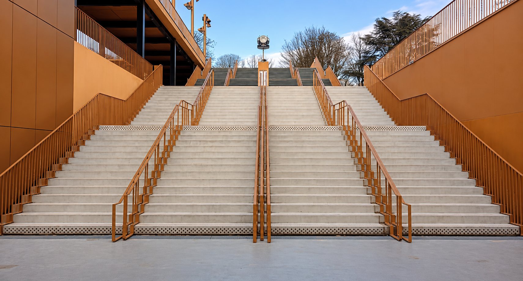 Un large escalier en béton clair divisé par plusieurs rampes métalliques dorées, encadré par de hauts murs orangés sous un ciel bleu.