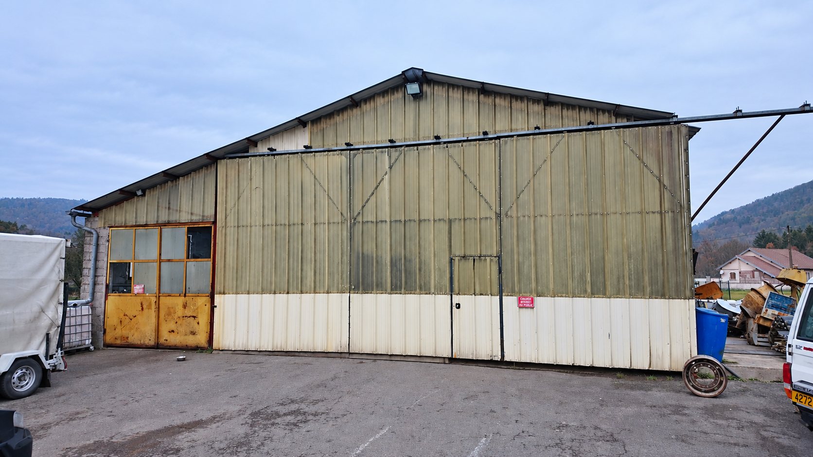 Un grand hangar métallique aux panneaux verdis et blancs, avec une porte jaune et une cour goudronnée, au pied de collines.