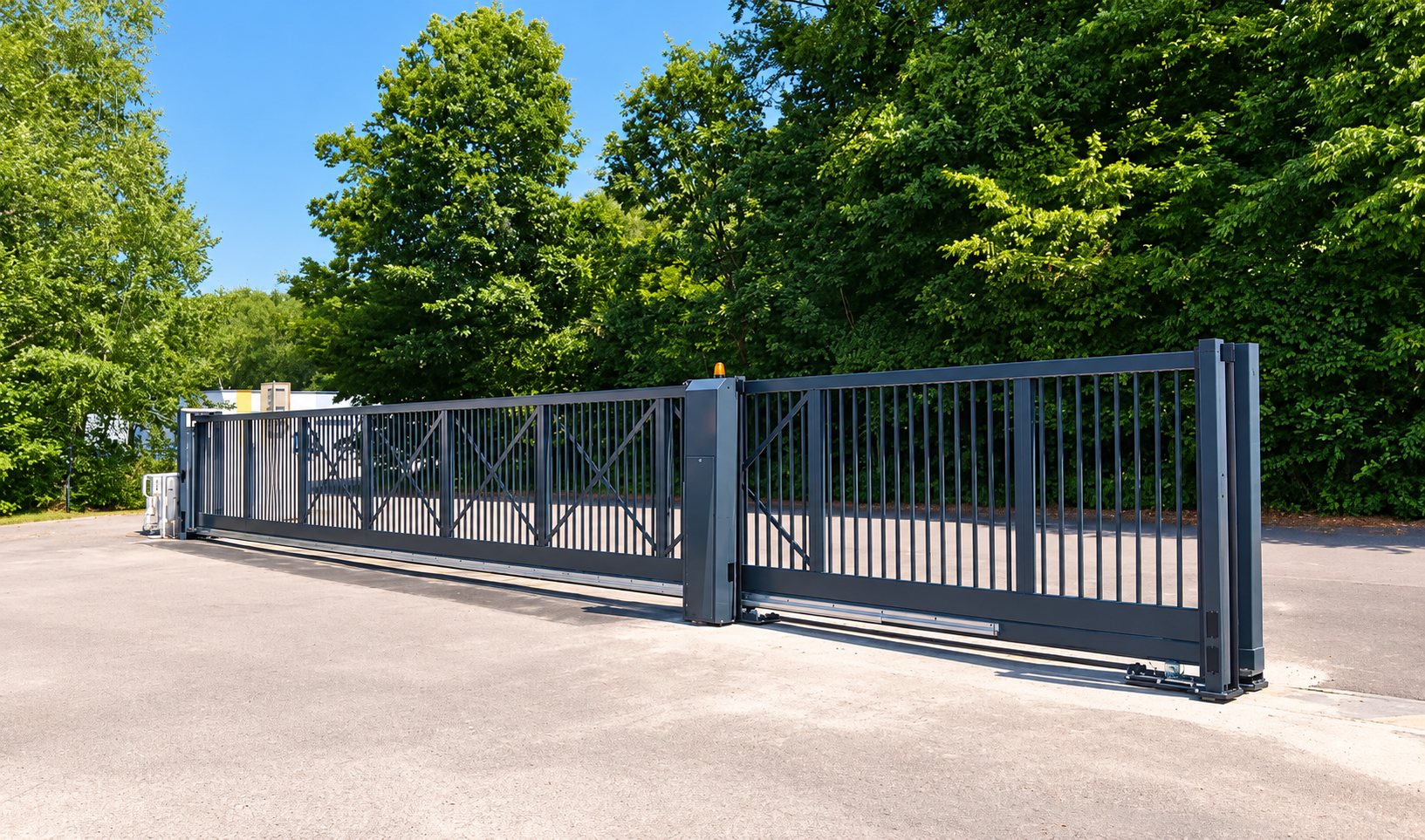 Un grand portail coulissant gris anthracite à barreaux verticaux, installé à l'entrée d'un parking bordé d'arbres sous un ciel bleu.