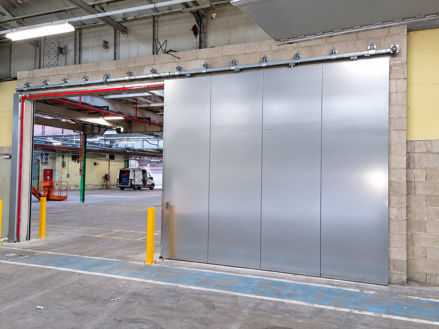Une grande porte coulissante en acier galvanisé suspendue à un rail, installée dans l'ouverture d'un entrepôt, avec une camionnette blanche visible au fond.