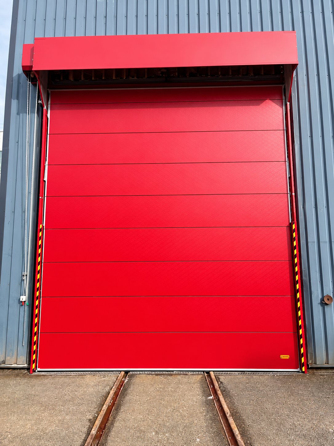 Une grande porte industrielle rouge à panneaux horizontaux, équipée de bandes de sécurité jaunes et noires, est installée sur la façade métallique grise d'un entrepôt, avec des rails au sol devant.