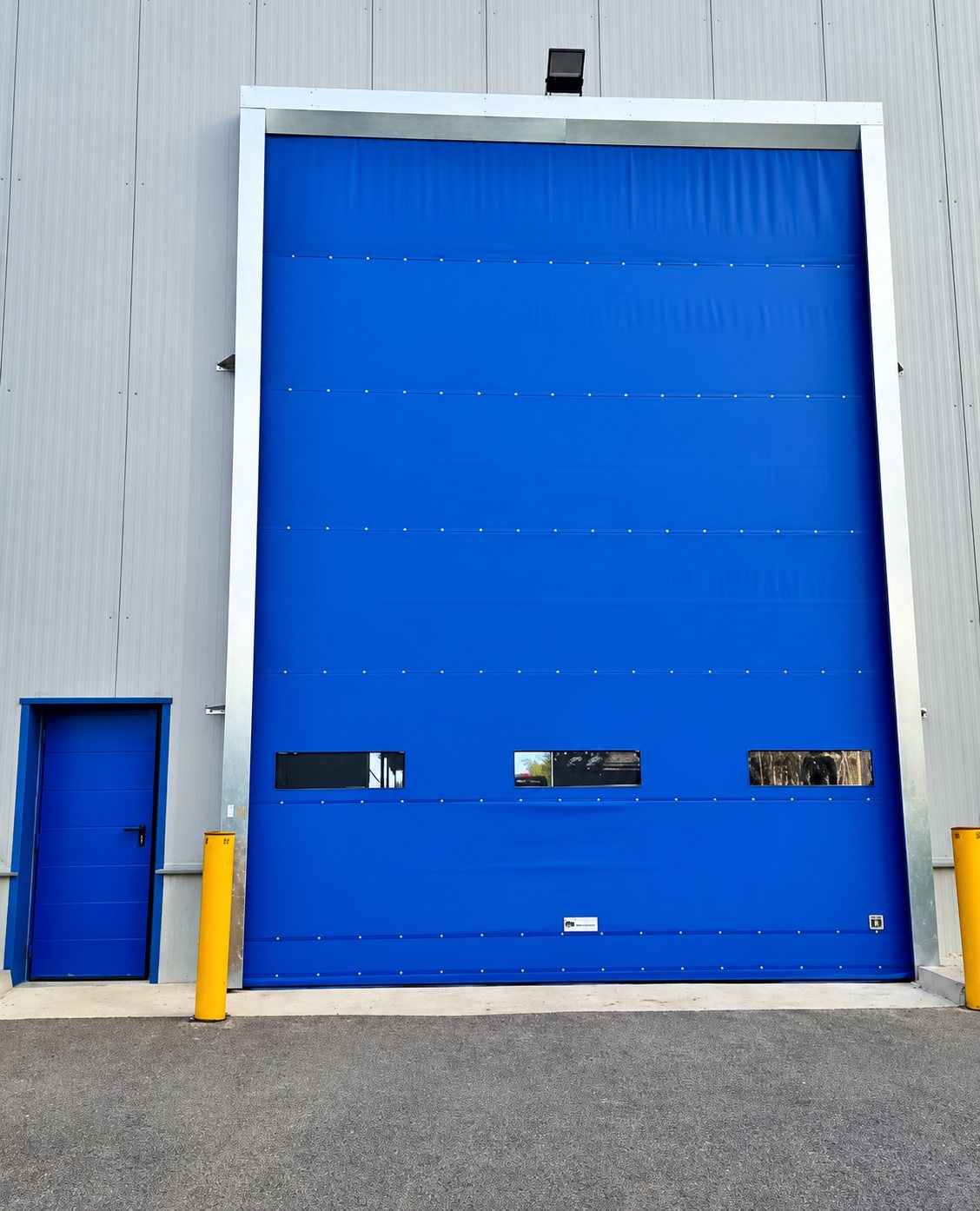 Une grande porte industrielle bleue à hublots et une petite porte bleue sont installées sur la façade d'un entrepôt gris, avec des poteaux de protection jaunes devant.