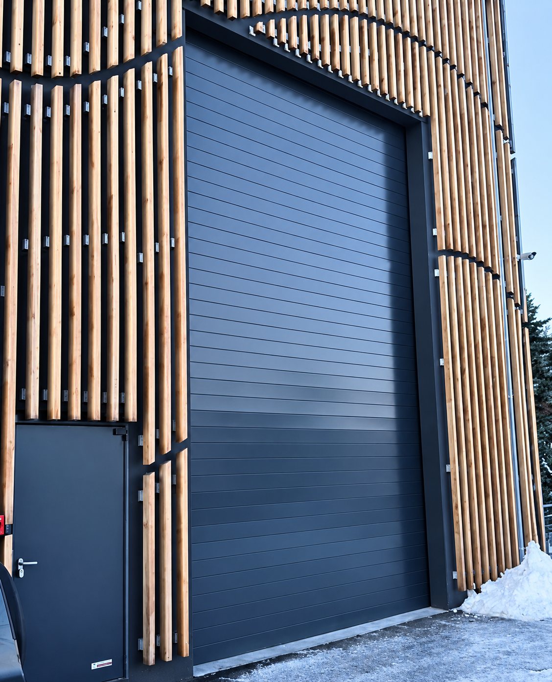 Une grande porte sectionnelle bleu foncé et une porte piétonne sont encadrées par une façade à lames de bois verticales, avec un peu de neige au sol.