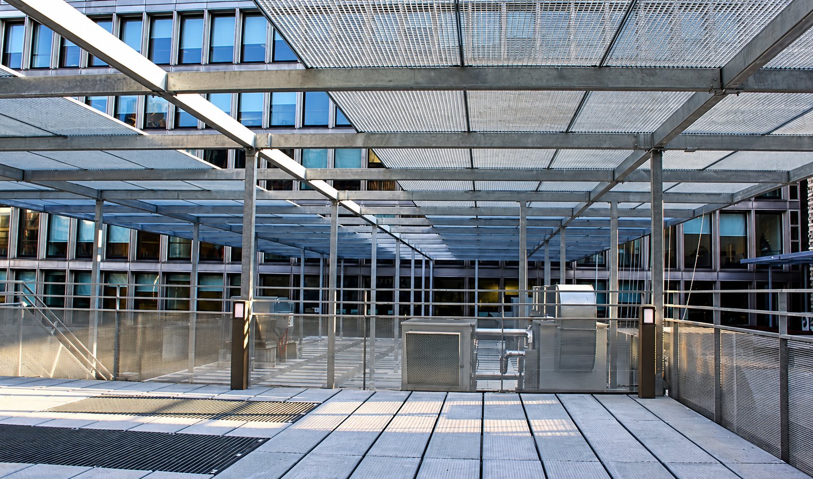 Une grande pergola en acier galvanisé avec toiture en caillebotis métallique couvre une terrasse dallée devant un immeuble de bureaux vitré.