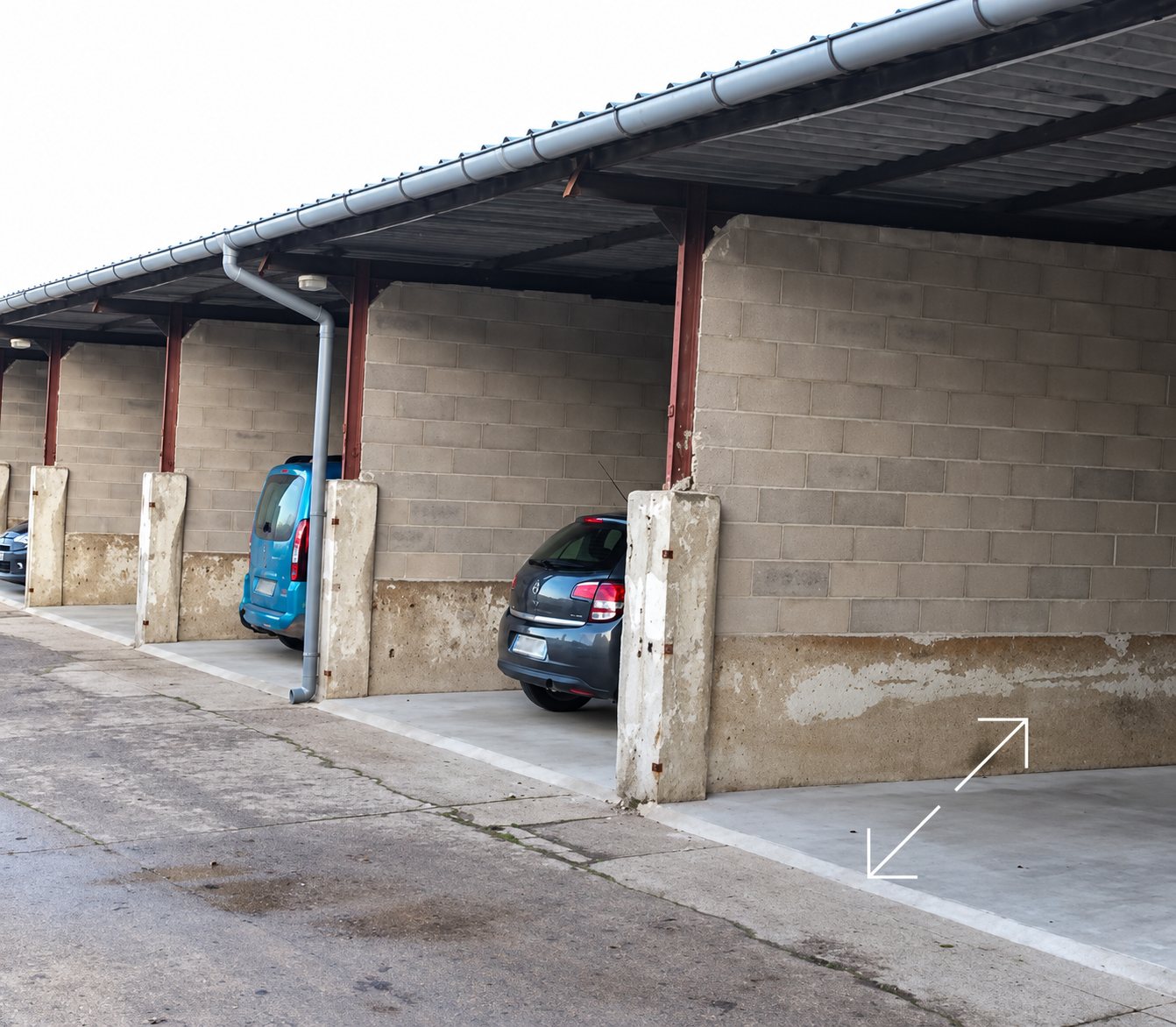 Des voitures sont garées dans une rangée de box ouverts à toit métallique, séparés par des murs en parpaings et des piliers en béton.