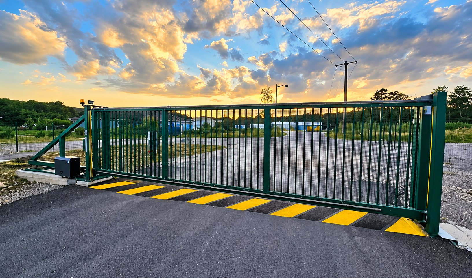 Un grand portail coulissant vert à barreaux, équipé d'un moteur et bordé de bandes jaunes au sol, ferme l'entrée d'un site industriel sous un ciel de coucher de soleil.