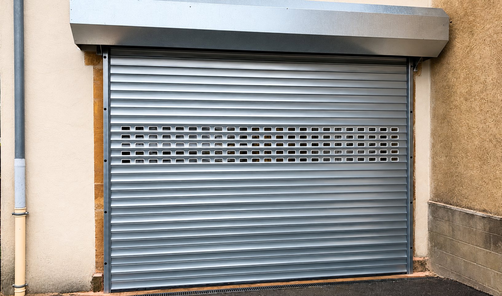 Un rideau métallique gris à lames avec une rangée d'ouvertures rectangulaires, posé sur la façade d'un bâtiment en pierre et crépi.