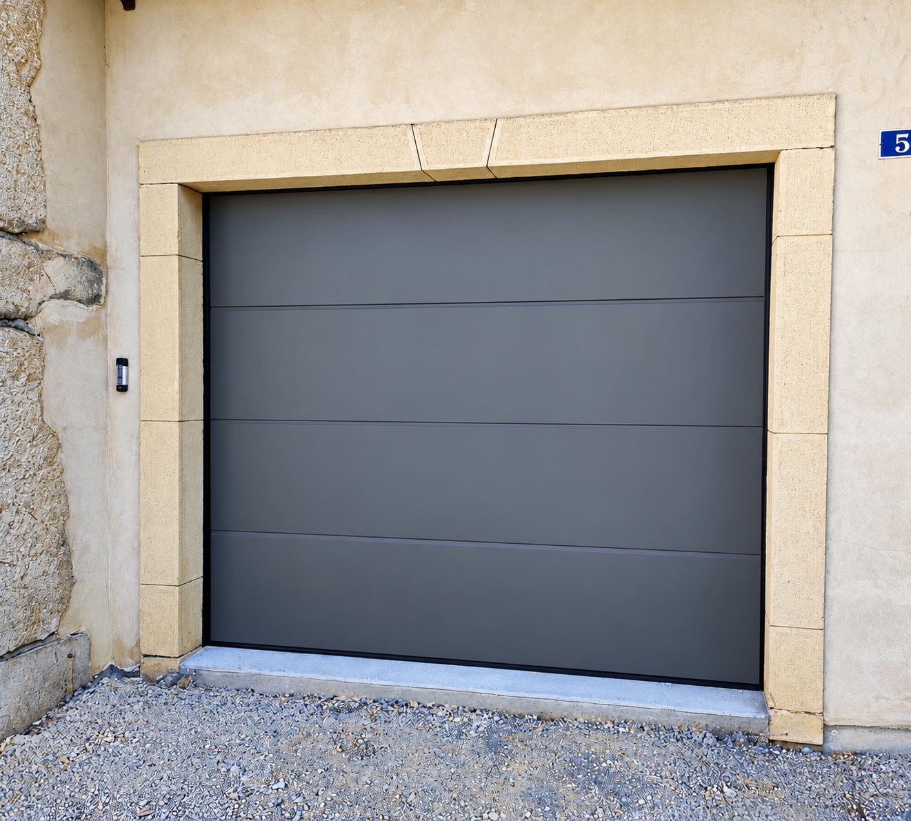 Porte de garage sectionnelle grise installée dans un encadrement en pierre jaune clair sur une façade en crépi, avec un numéro de rue bleu à droite.
