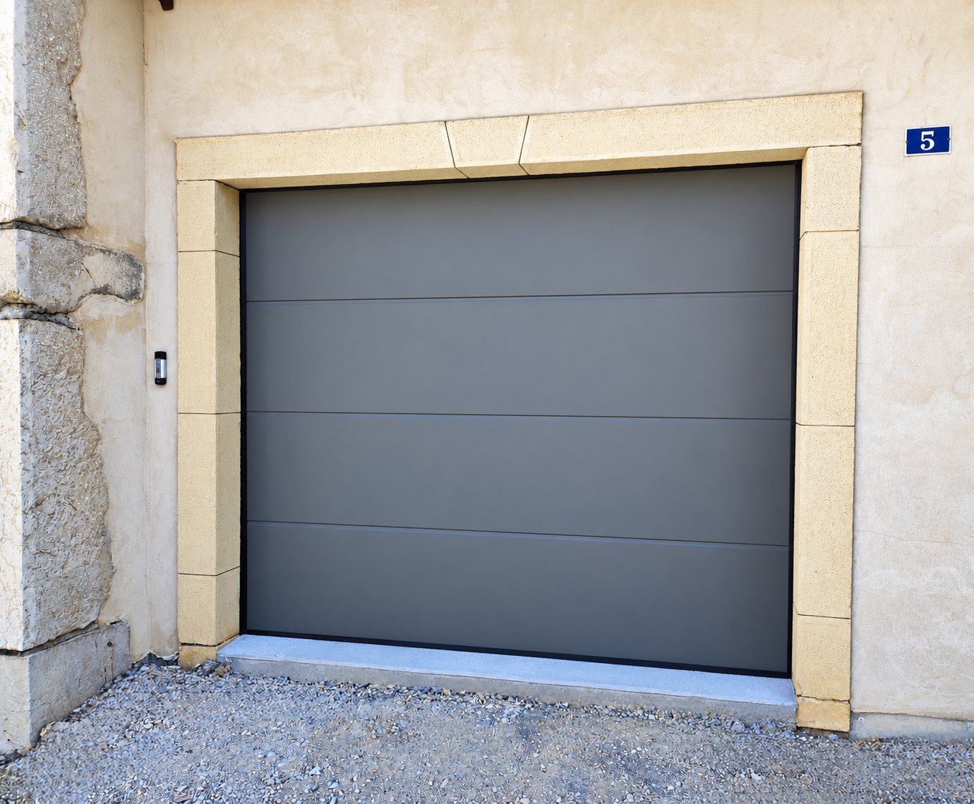 Porte de garage sectionnelle grise à panneaux lisses posée dans un encadrement en pierre claire, sur une façade en crépi portant le numéro 5.