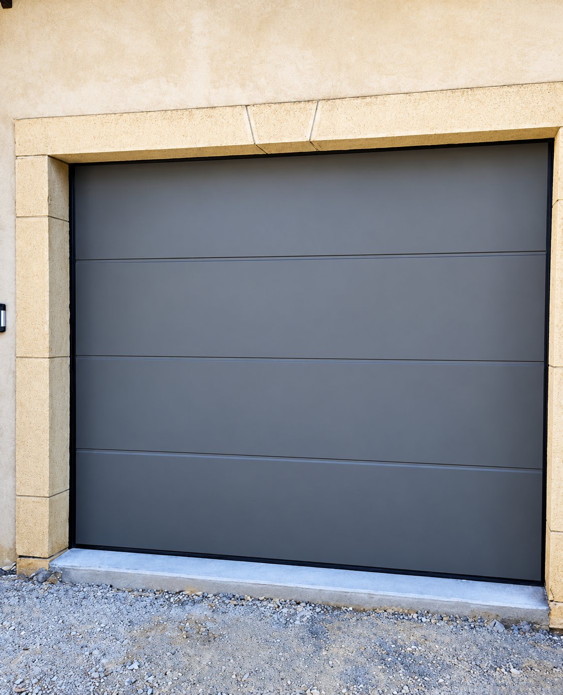 Une porte de garage sectionnelle grise à panneaux lisses est posée dans un encadrement de pierre beige, au-dessus d'un seuil en béton et d'un sol de gravier.