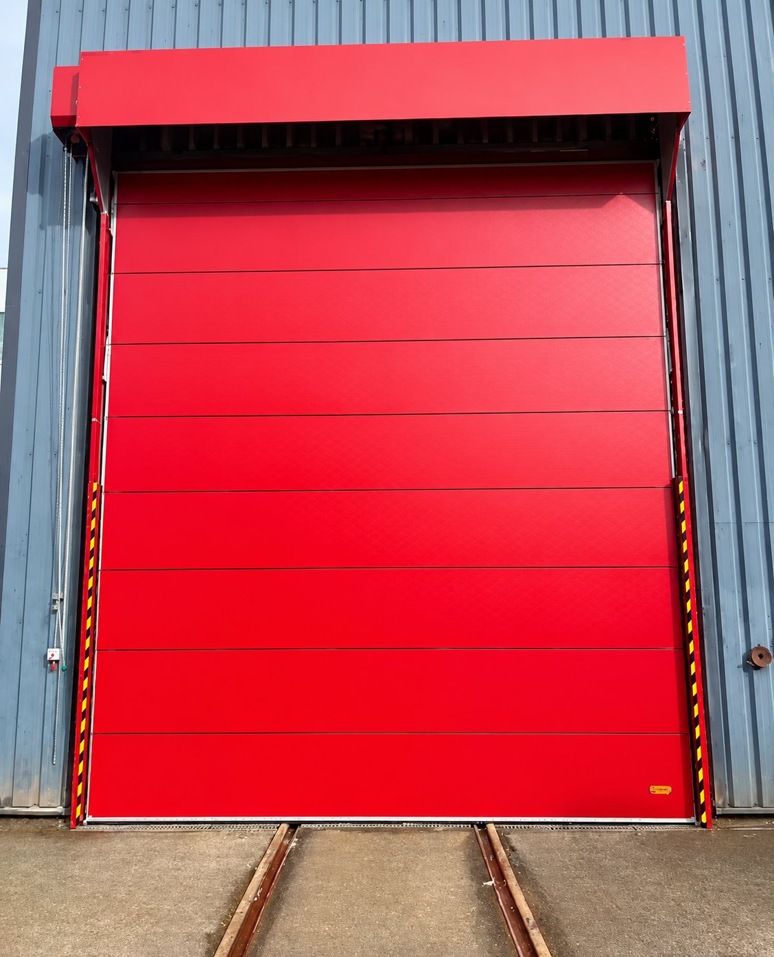 Une grande porte sectionnelle industrielle rouge installée sur la façade d'un entrepôt en bardage gris, avec des rails au sol devant l'entrée.