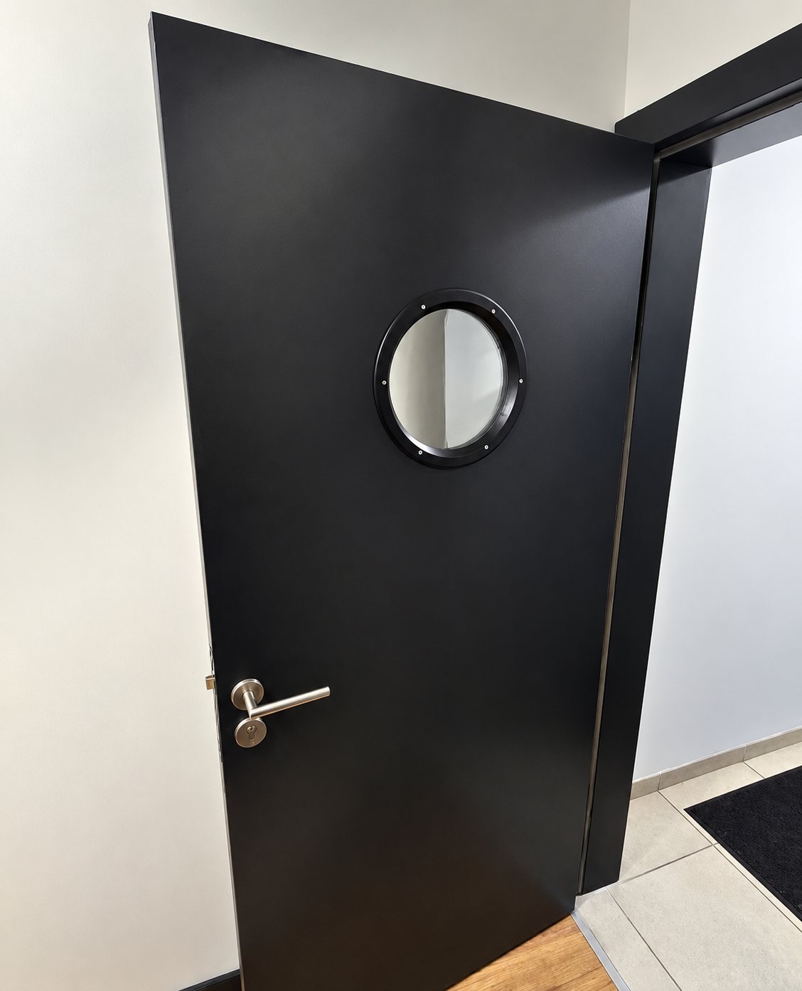 Une porte intérieure noire ouverte, équipée d'un petit hublot rond et d'une poignée en inox, dans un couloir aux murs clairs.