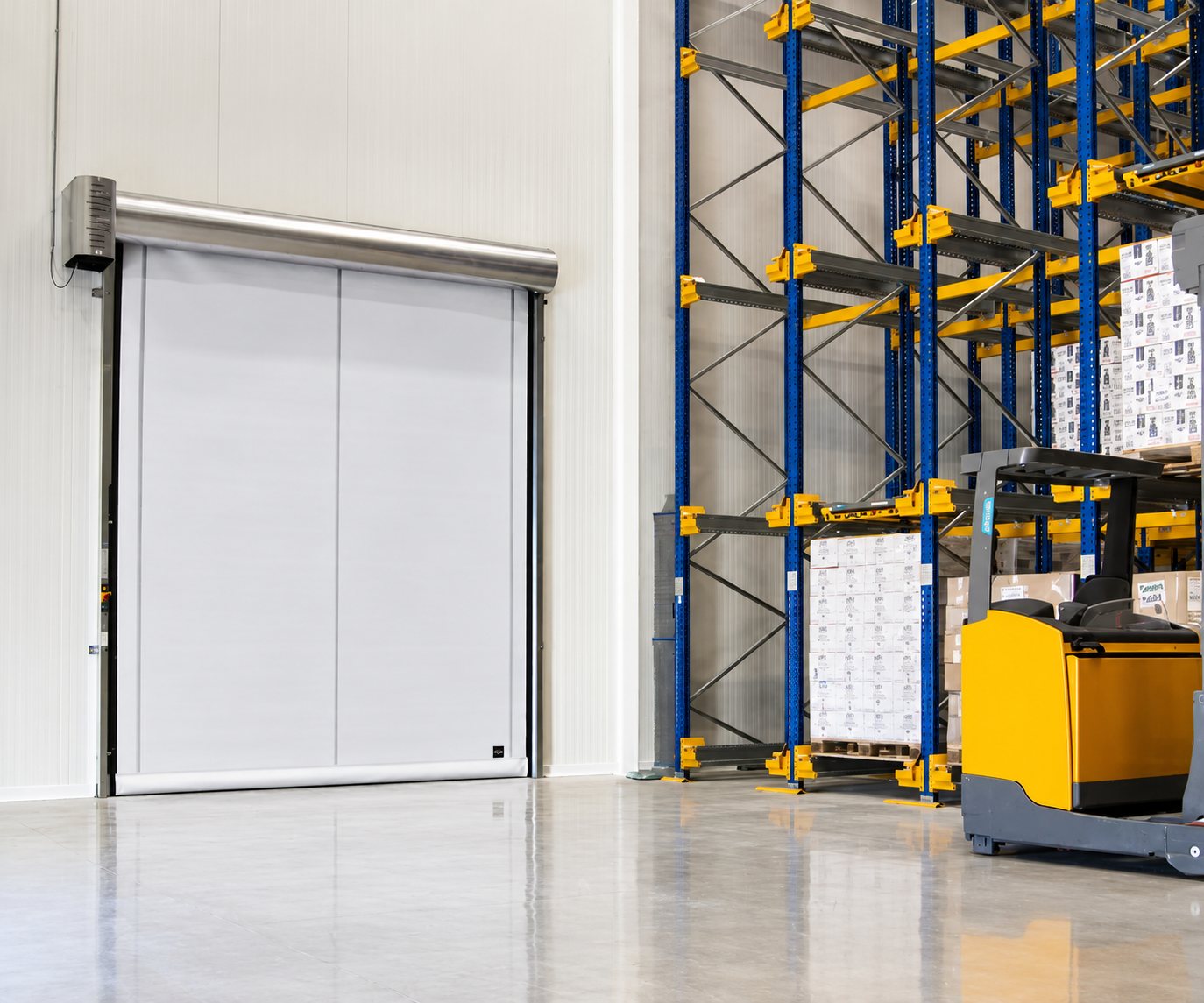 Une porte rapide enroulable grise installée dans un entrepôt, à côté de rayonnages bleus chargés de cartons et d'un chariot élévateur jaune.