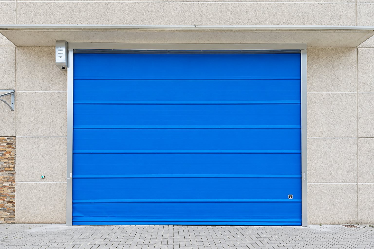 Une grande porte rapide en tissu bleu, encadrée d'aluminium, est installée dans un mur de béton clair au-dessus d'un sol pavé.
