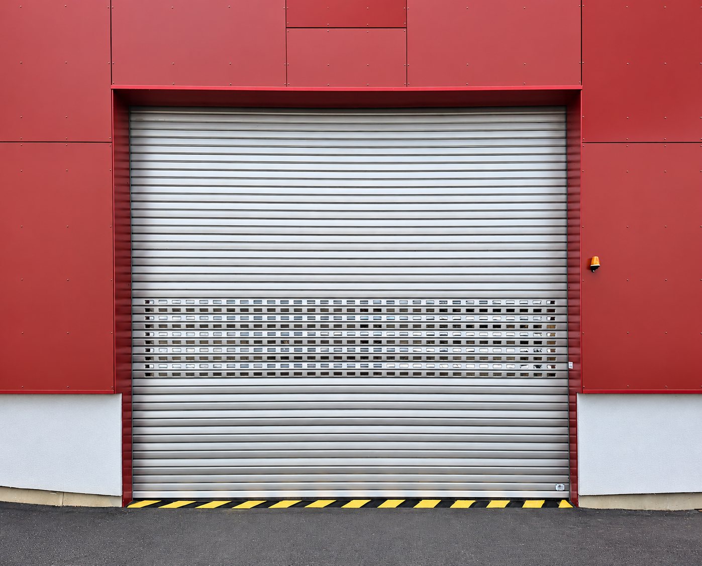 Rideau métallique en aluminium avec bandeau micro-perforé, installé dans une façade de bardage rouge, avec marquage jaune et noir au sol et feu orange de signalisation.