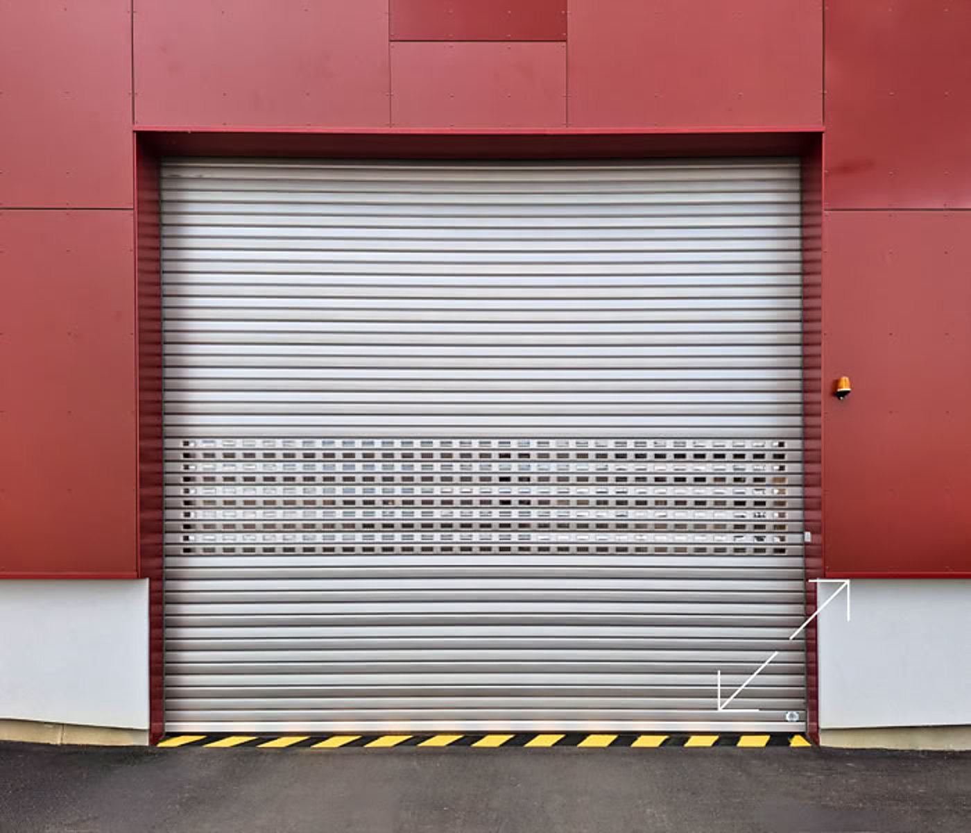 Une grande porte roulante en aluminium avec une partie perforée, installée dans une façade de bardage rouge, avec une bande de sécurité jaune et noire au sol.