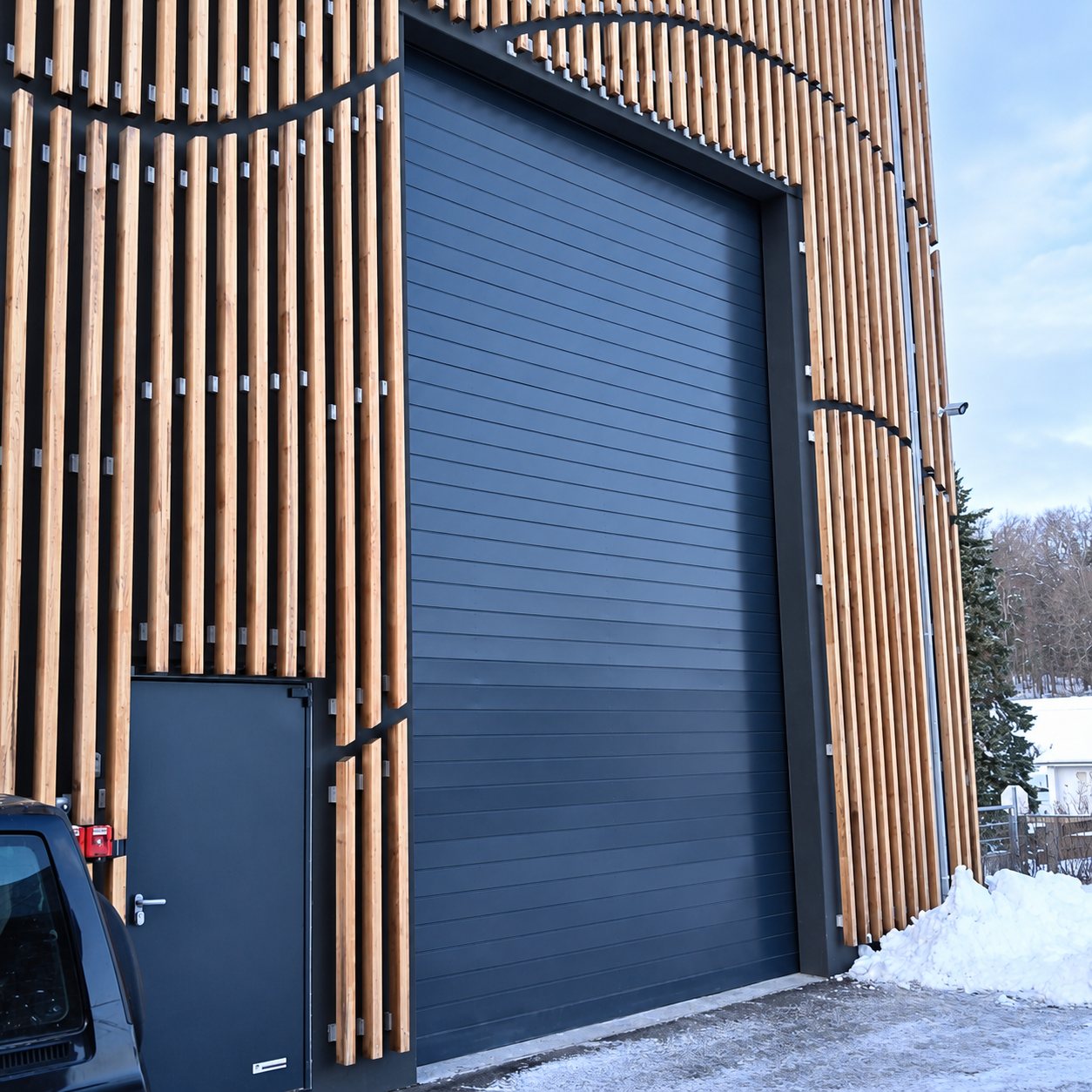 Une grande porte sectionnelle grise et une porte piétonne s'intègrent dans une façade à claire-voie en lattes de bois, avec de la neige au sol.