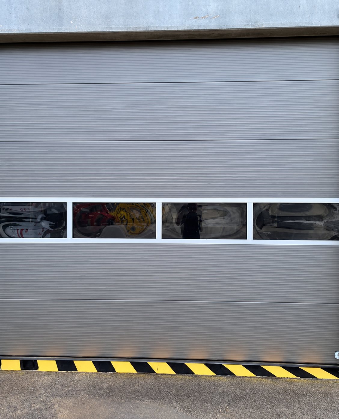 Porte sectionnelle industrielle grise équipée d'une rangée de quatre hublots vitrés et d'une bande de sécurité jaune et noire au sol.