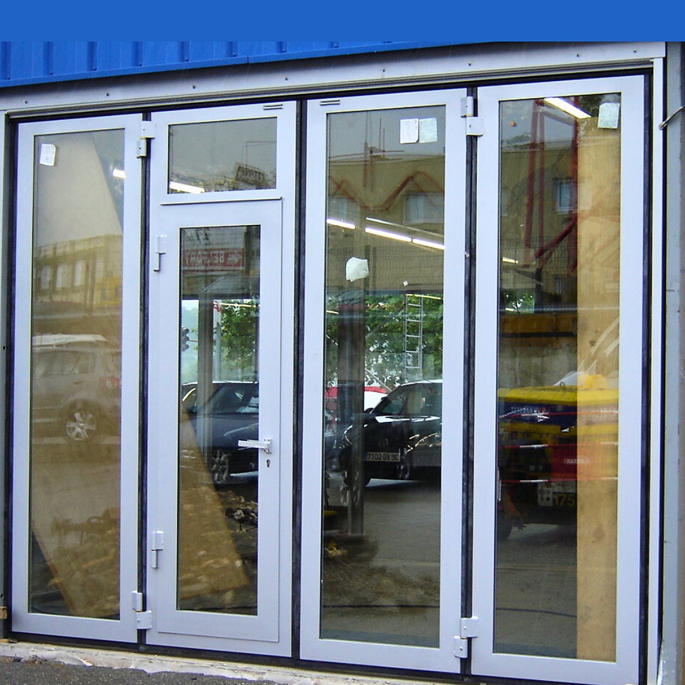 Une porte pliante vitrée à cadre aluminium blanc, avec une porte d'accès intégrée, installée dans un bâtiment à bardage bleu et reflétant des voitures et des arbres.