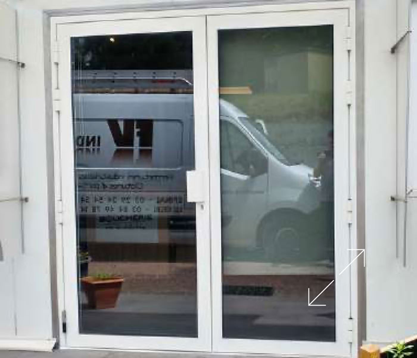 Porte vitrée à double battant avec un cadre blanc, dans laquelle se reflète une camionnette blanche garée dans la rue.