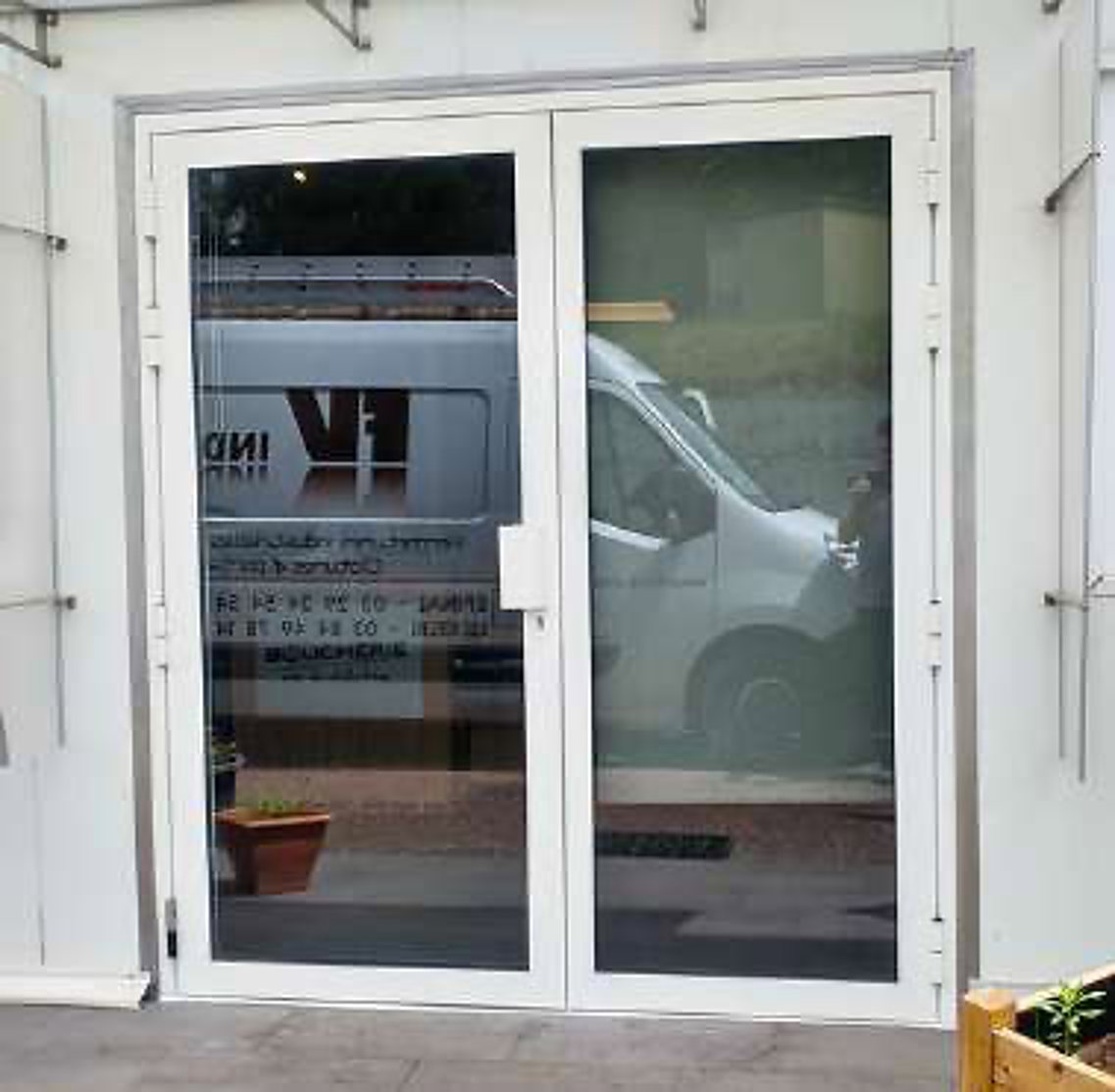 Une porte vitrée à double battant à cadre blanc dans laquelle se reflète une camionnette blanche garée devant le bâtiment.