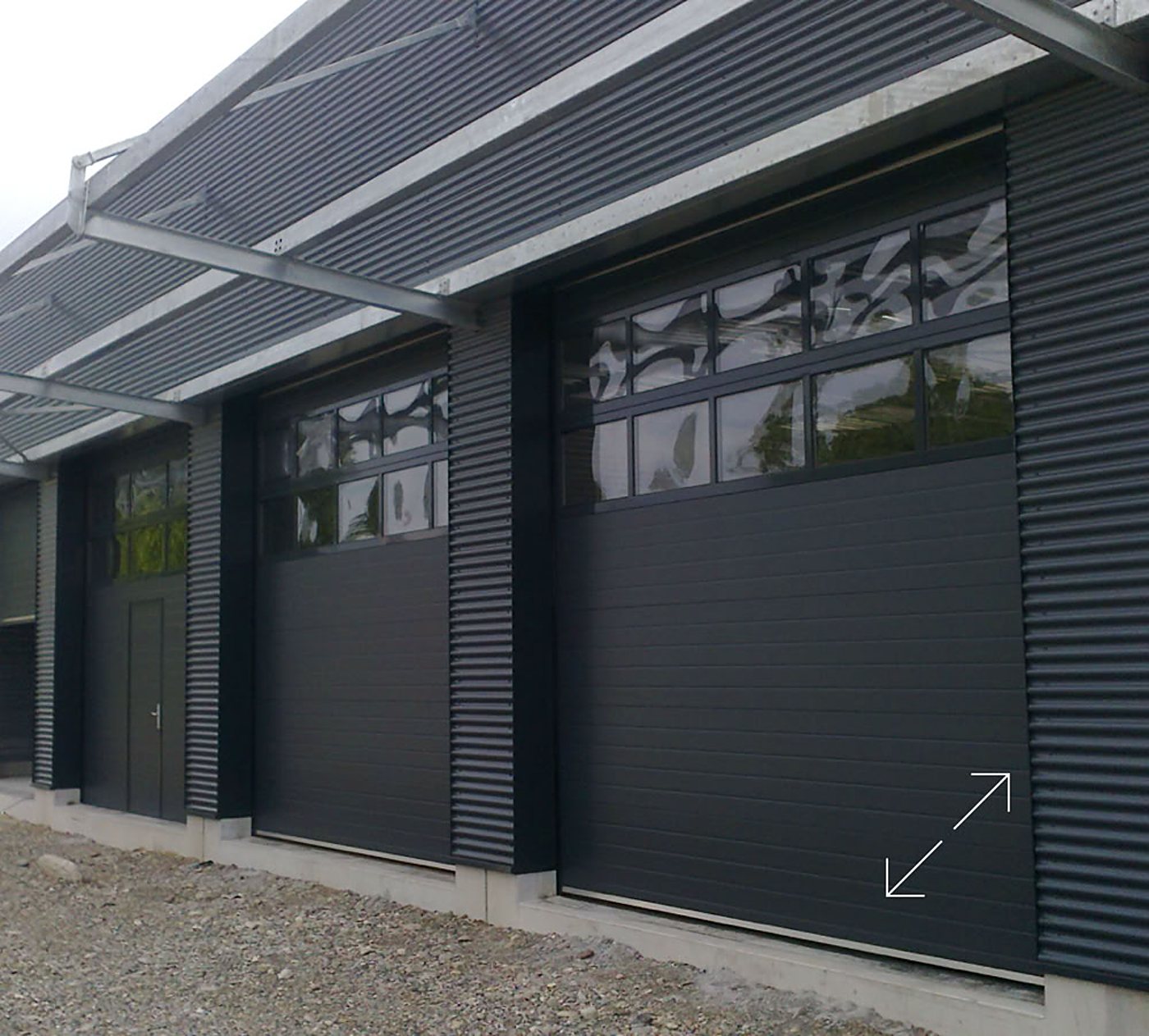 Façade d'un bâtiment industriel en tôle ondulée sombre équipé de deux grandes portes sectionnelles anthracite vitrées et d'une porte piétonne.