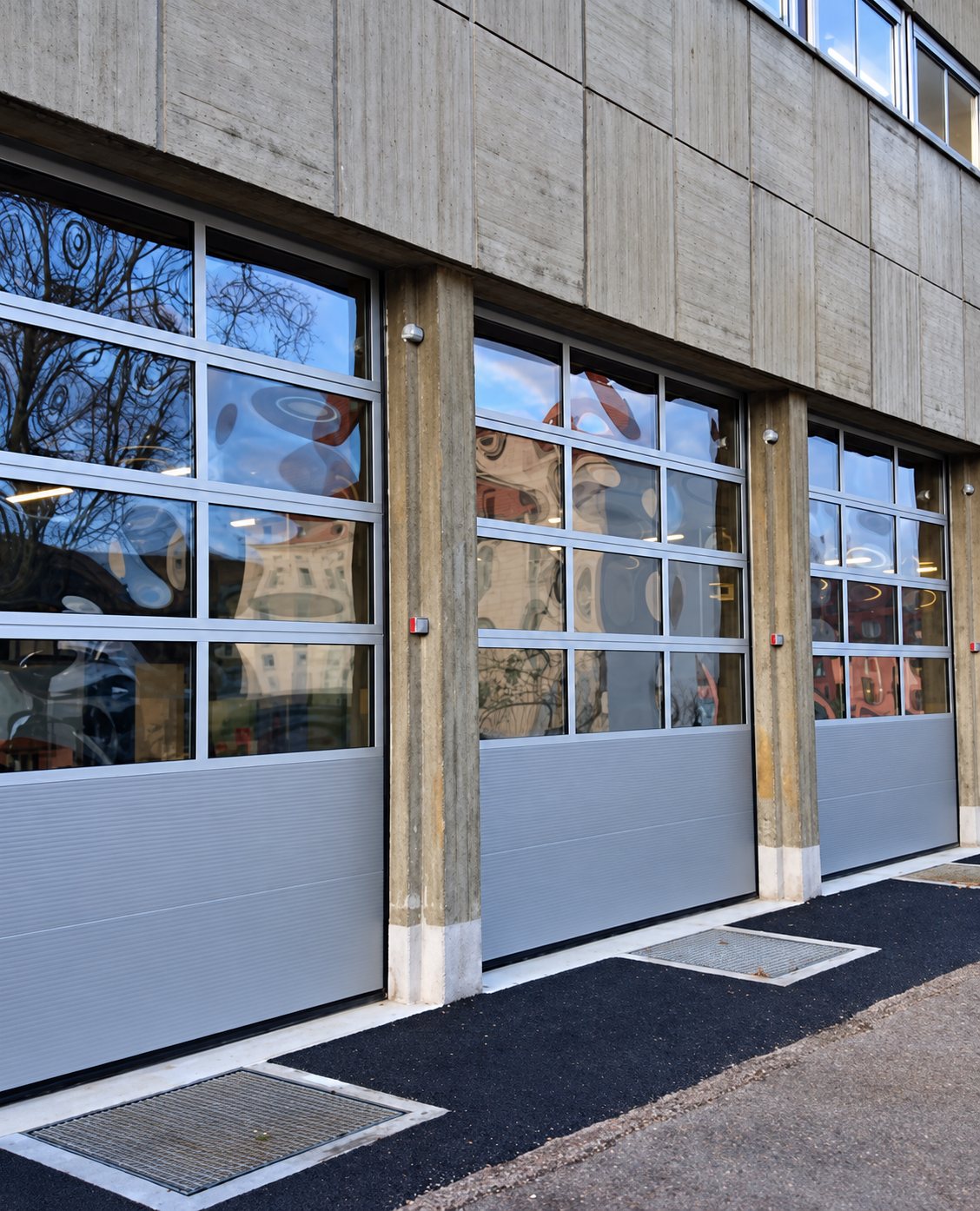 Trois grandes portes sectionnelles en aluminium avec partie vitrée, installées dans la façade en béton d'un bâtiment, au-dessus d'un sol en enrobé noir.