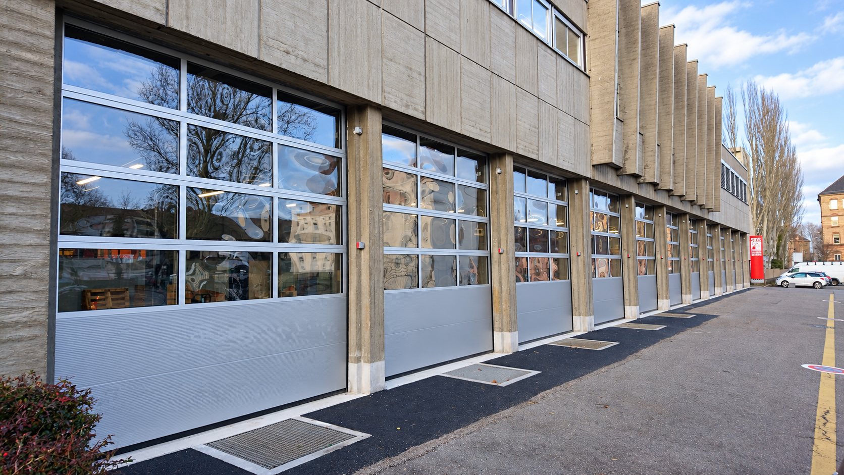 Une longue rangée de grandes portes sectionnelles en aluminium gris avec partie vitrée, installées sur la façade en béton d'un bâtiment public.
