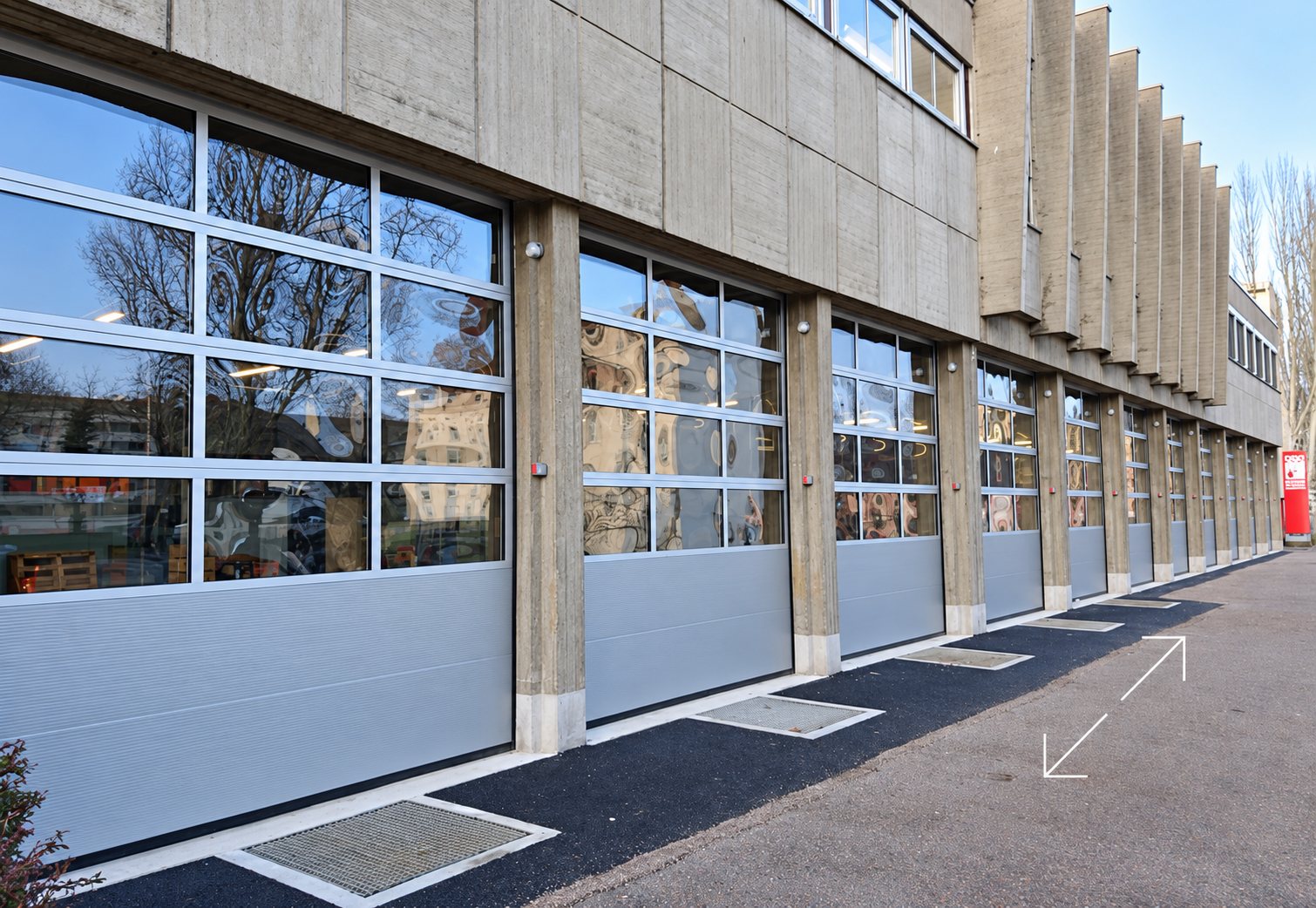 Une rangée de portes sectionnelles grises à panneaux vitrés installées sur la façade en béton d'un bâtiment, avec un sol en enrobé noir devant.