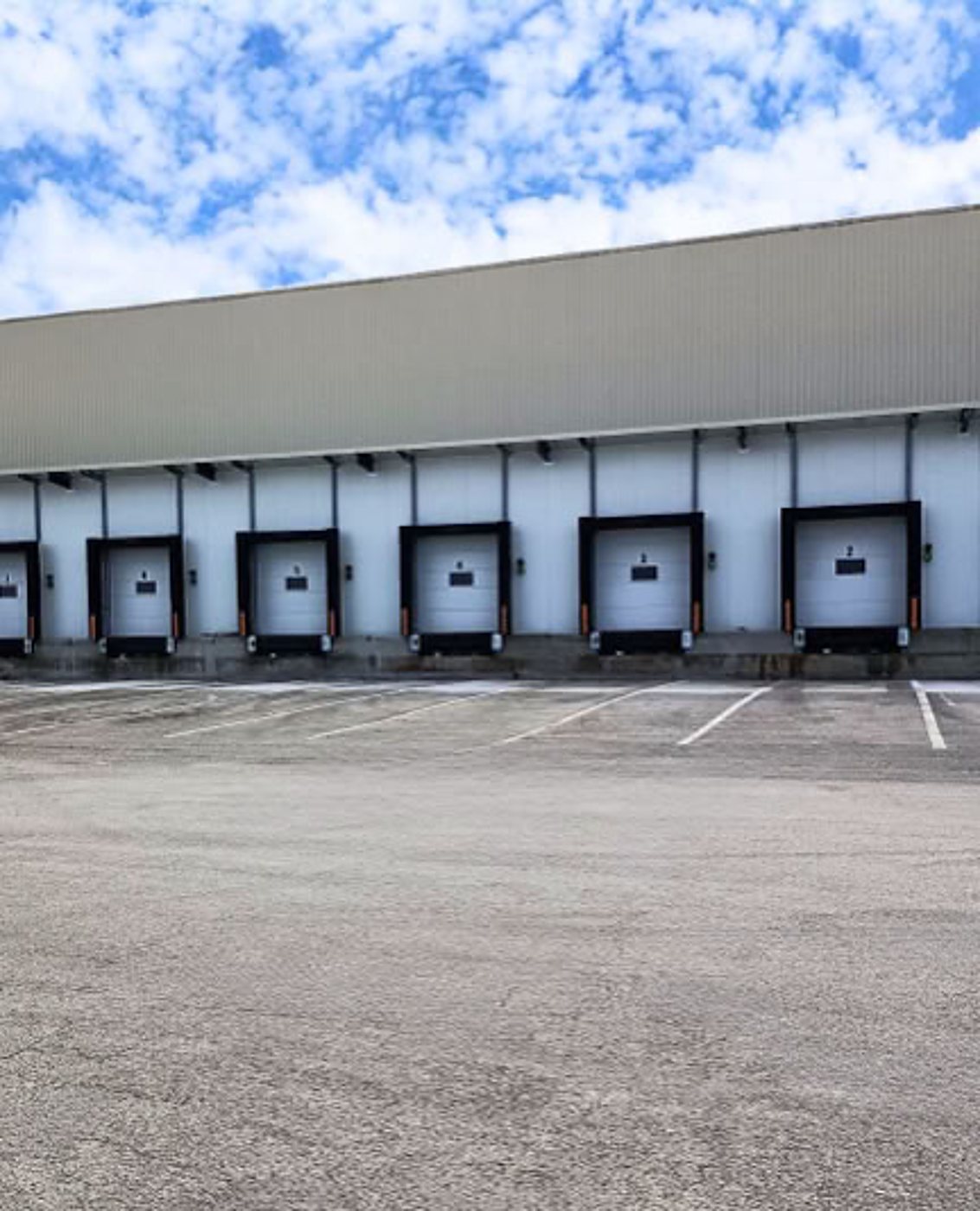 Façade d'un entrepôt logistique avec une rangée de quais de chargement aux portes grises, devant un grand parking vide sous un ciel nuageux.