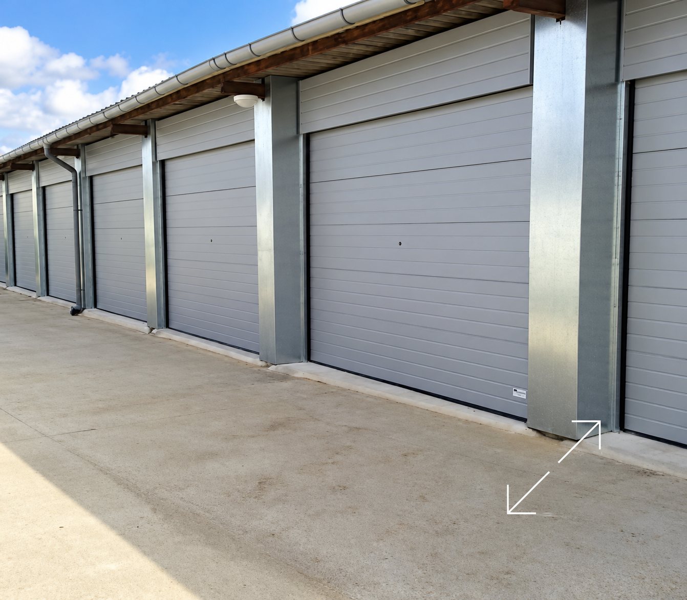 Une rangée de garages à portes sectionnelles grises, séparées par des poteaux métalliques, devant une dalle en béton sous un ciel bleu.